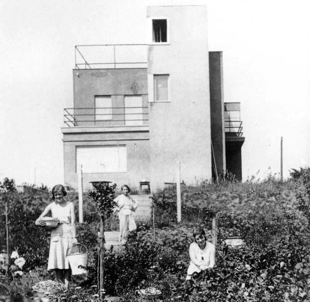 Die Fotografie aus dem Jahr 1930 zeigt drei in luftigem Weiß gekleidete Frauen inmitten eines üppigen sommerlichen Gartens. Die beiden jungen Frauen im Vordergrund blicken von der Ernte auf und lächeln in die Kamera. Hinter ihnen führen Treppen einen kleinen Hügel hinauf, hin zu einem Haus mit klaren und einfachen Linien. Auf einer dieser Treppen steht die Malerin Lucy Ortlepp, selbstbewusst und entspannt blickt sie dem Fotografen entgegen – es ist ihr neues Zuhause, vor dem sie zu sehen ist. Das Haus befindet sich am Weimarer Ratstannenweg 21. Noch heute ist das Namensschild der Ortlepps an seiner Eingangspforte angebracht – in Erinnerung an die früheren Besitzer.